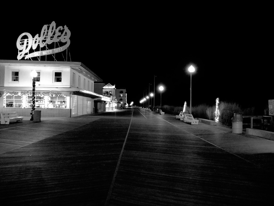 An empty boardwalk at night, street lamps along the beach edge, a large sign above a two-story building sign reads 'Dolles.'