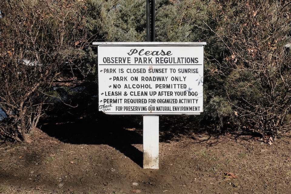 A small rectangular, wooden sign on a wooden post. The white paint is beginning to chip, the black letters are faded. The words appear hand stenciled, except for wide script of 'please' and 'thank you.' The sign reads: 'Please, observe park regulations. Park is closed sunset to sunrise. Park on roadway only. No alcohol permitted. Leash and clean up after your dog. Permit required for organized activity. Thank you for preserving our natural environment.'