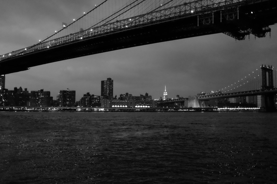 A clear night sky, the view from a pier under the Brooklyn Bridge looking north and across the East River. The Manhattan Bridge in the distance and behind it the bright white lights of The Empire State building.