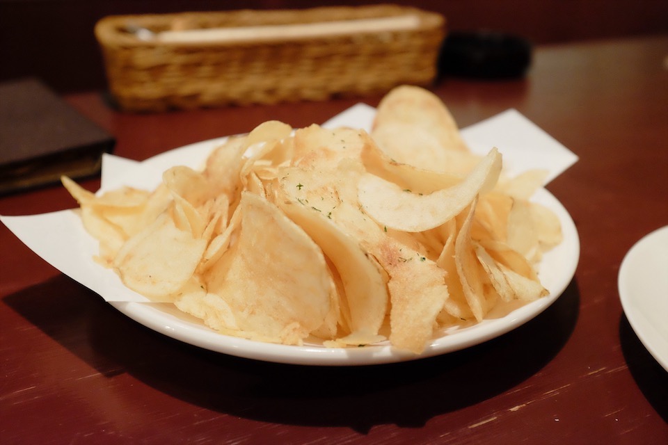 Golden yellow potato chips, dusted with bright green flecks of dill, sit on a white plate that rests on a wooden bar.