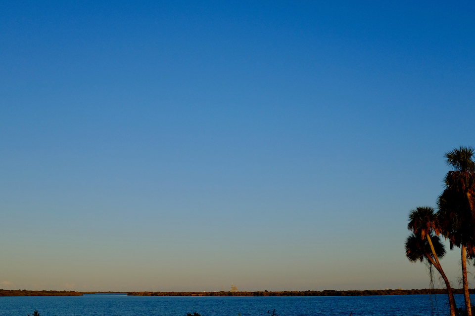 “Palm trees and water, looking toward Cape Canaveral in the distance where a rocket, not destined to launch this day, sits waiting in front of a blue sky, beginning to turn pale orange in the fading light.”