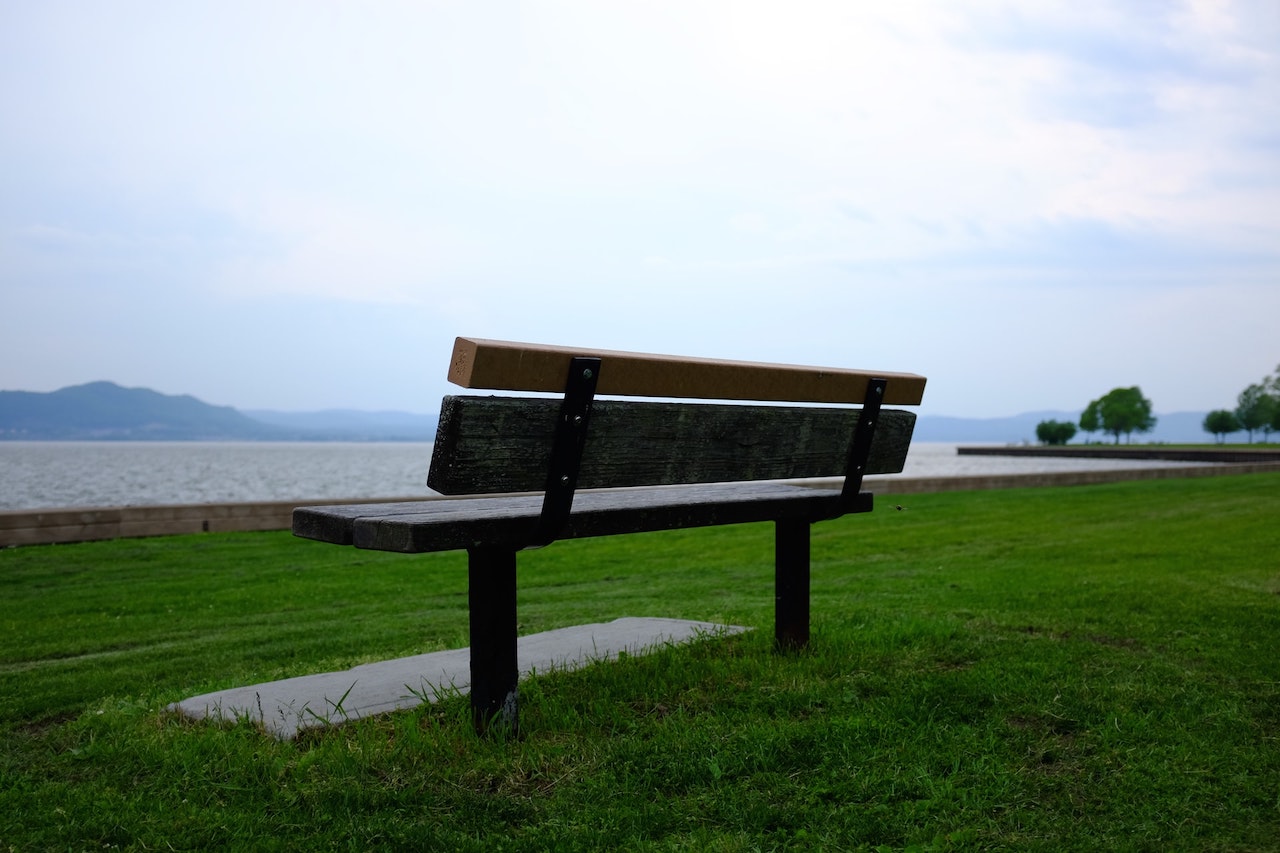 A wooden bench, sitting on green grass, overlooking the Hudson River in Croton Point Park, NY under a bright, pale, blue sky.